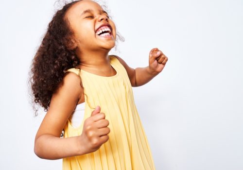 Portrait of African-American little curly haired girl in yellow dress laughing and dancing with closed eyes against white background